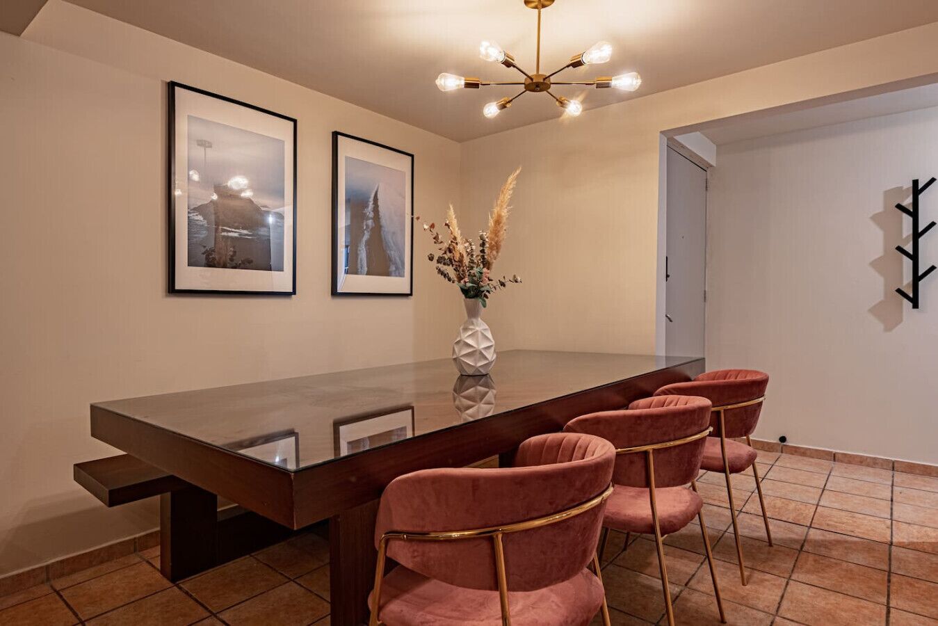 A dining area with a long table, four chairs, a vase with dried plants, and two framed pictures on the wall.