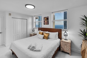 Ein Schlafzimmer mit einem großen Bett, weißer Bettwäsche, einem Holz-Kopfteil, einem Nachttisch mit Lampe, einem Fenster mit Blick auf Gebäude und das Meer sowie einer Tür links.