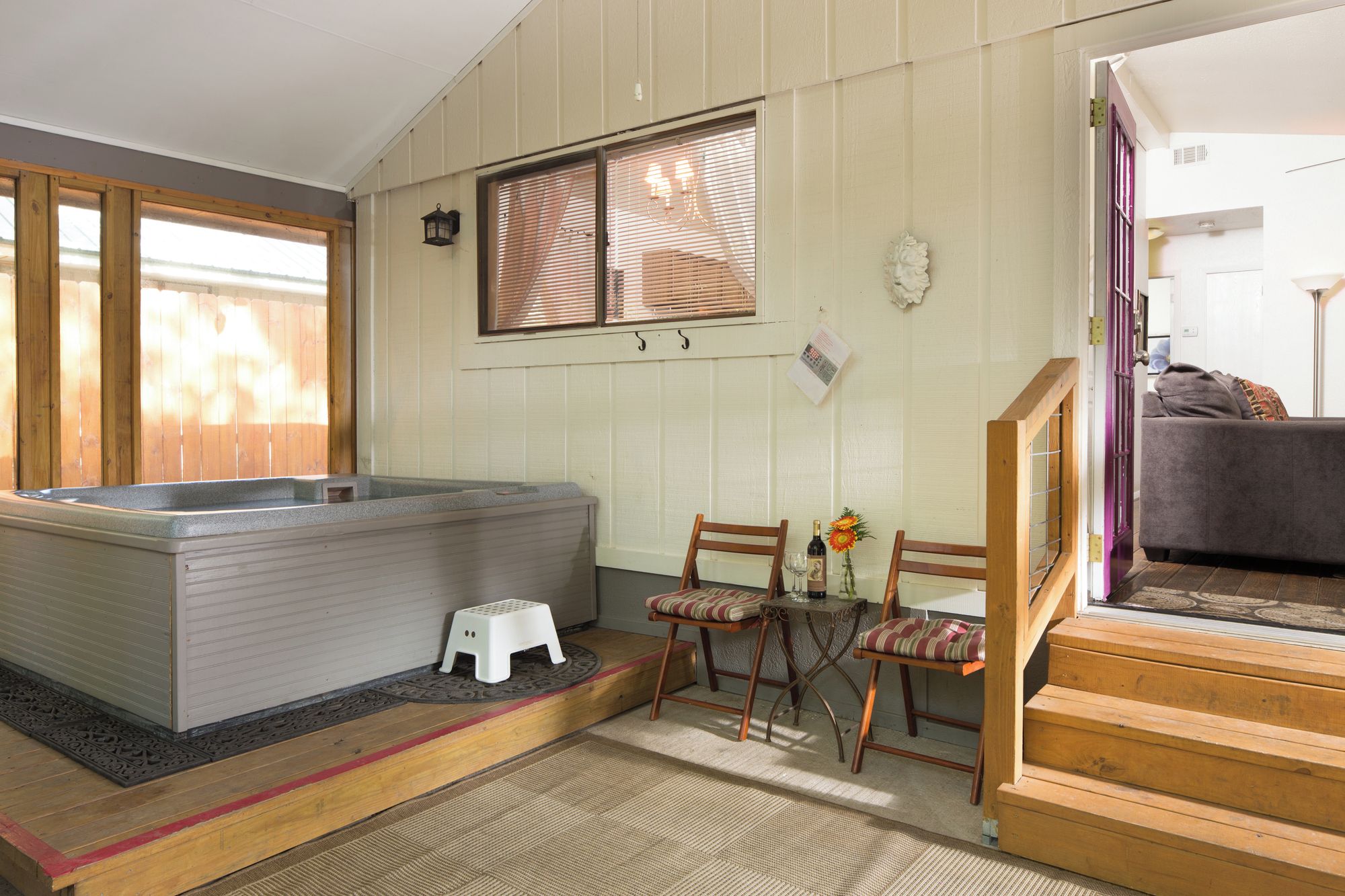 A room with a hot tub, wooden furniture, a small table with a bottle and flowers, and a staircase leading to another area.