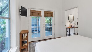 A bedroom with a large bed, a wooden nightstand, a TV mounted on the wall, a door with glass panels, and a small table with a mirror and a framed picture. - Discover Affordable Luxury in the Heart of Texas Hill Country (Wimberley)