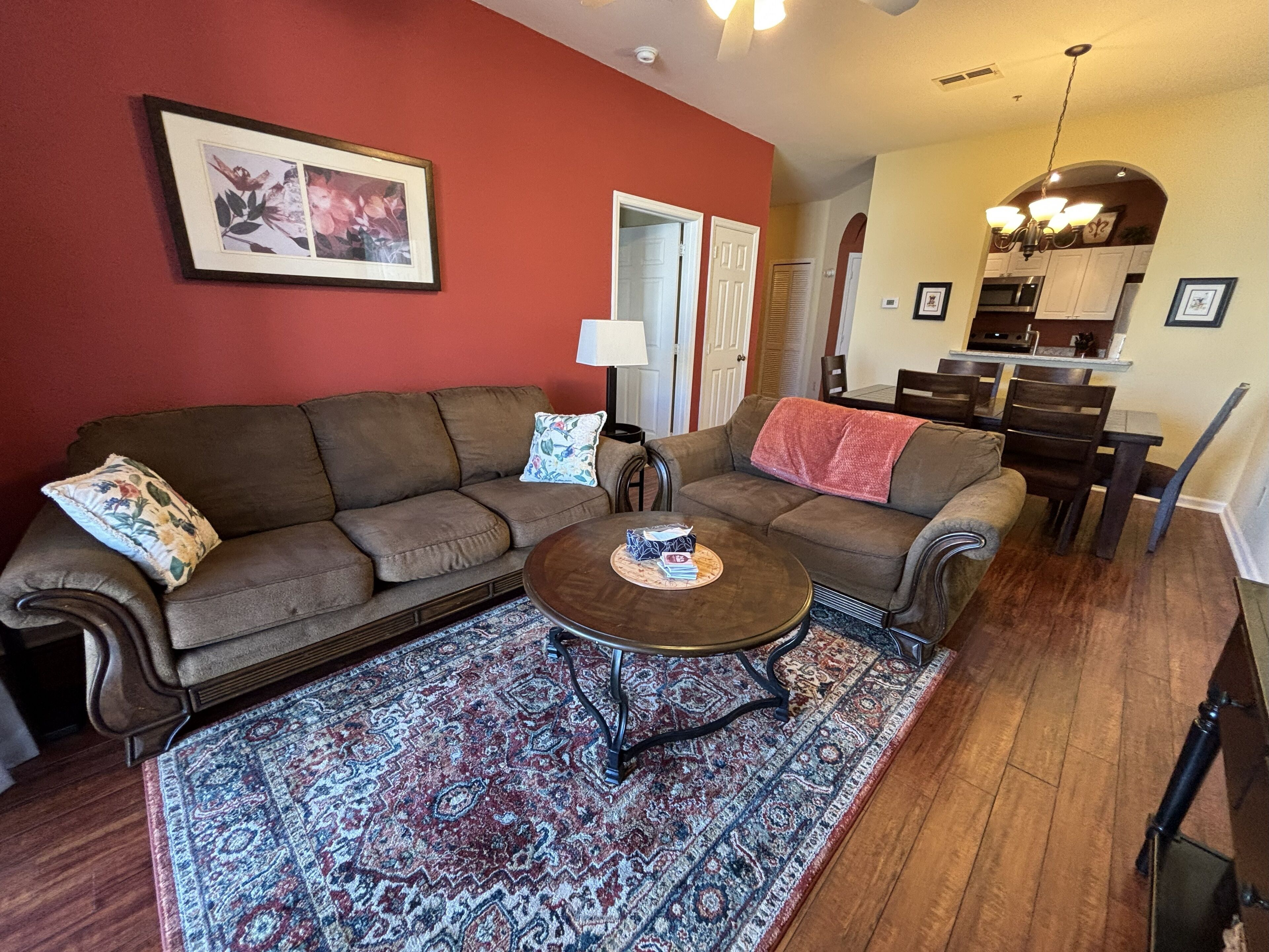A living room with a brown sofa, a coffee table, and a patterned rug. There is a kitchen visible in the background.