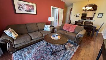 A living room with a brown sofa, a coffee table, and a patterned rug. There is a kitchen visible in the background.