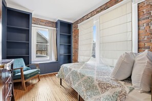 Een slaapkamer met een bed, een stoel, een raam en een bakstenen muur.
