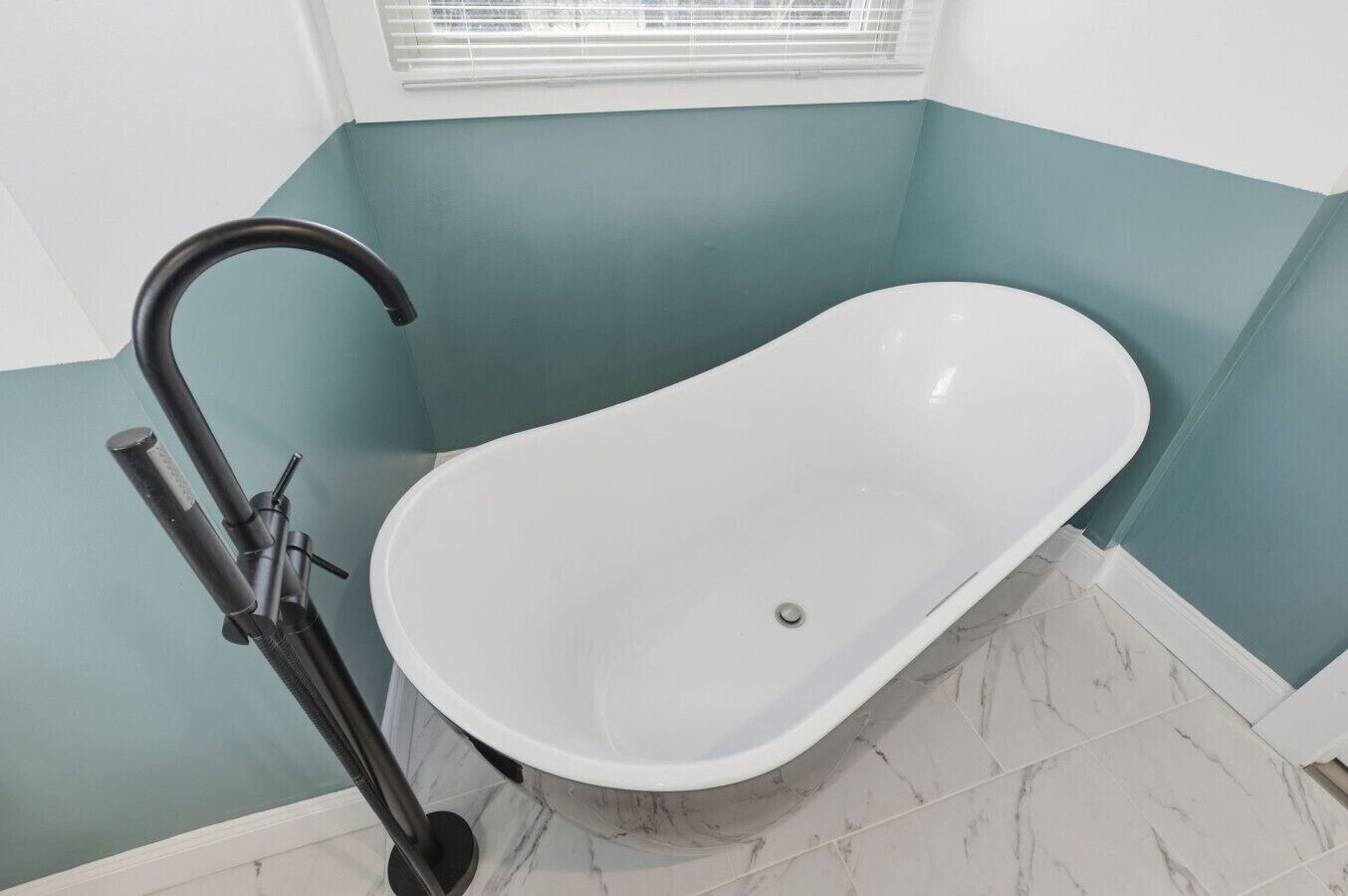 A bathroom with a freestanding bathtub, a handheld showerhead, and a window with blinds.