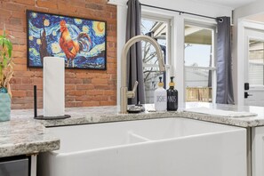 Een keuken met een granieten aanrecht, een roestvrijstalen spoelbak, een schilderij van een haan en een raam met gordijnen.