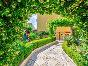 A paved pathway flanked by neatly trimmed hedges and flowering plants, leading to a gated entrance. - CIRO CH / HR-07098-04 by Interhome (Selce)