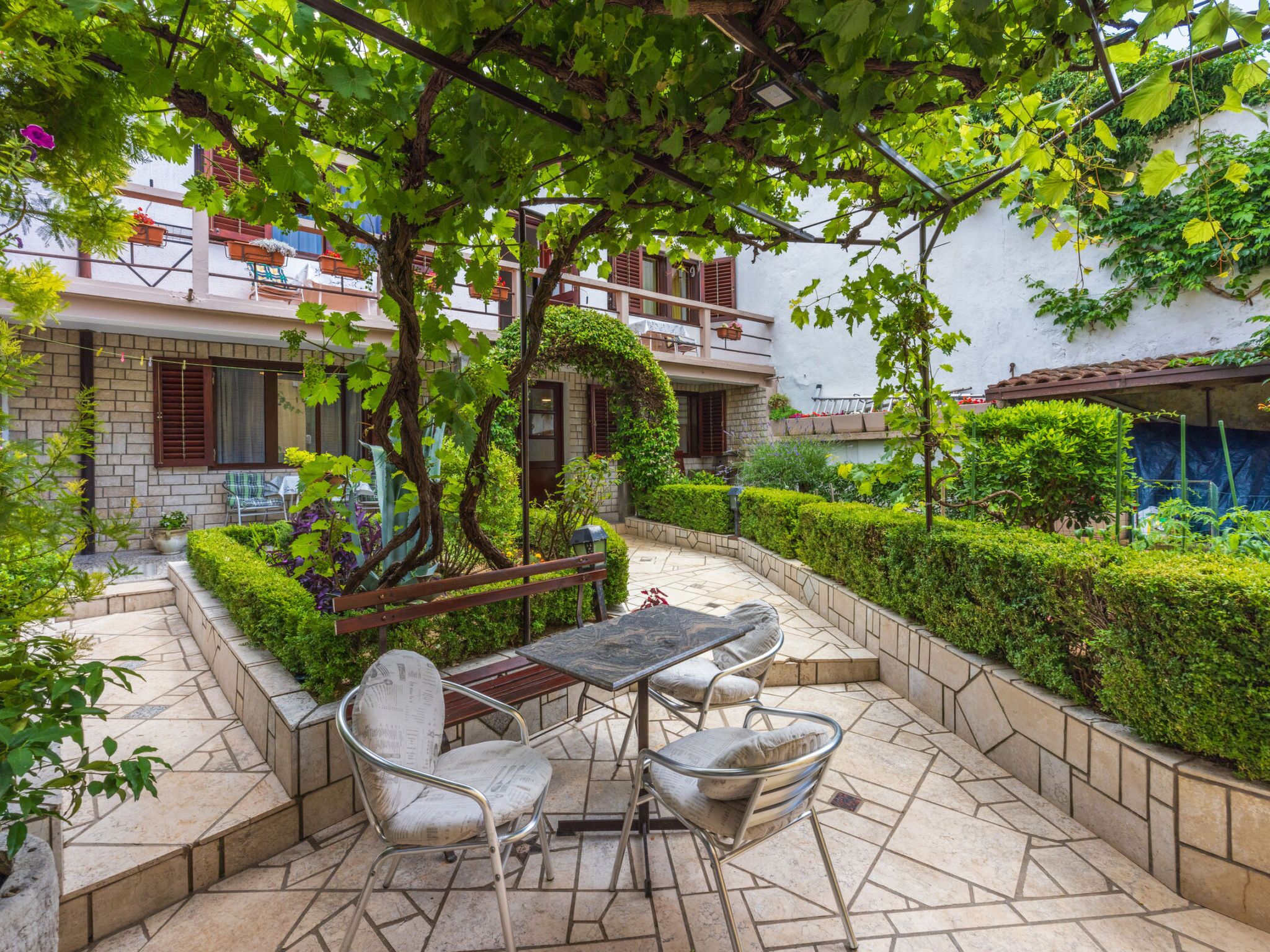 Une terrasse avec une table et des chaises, entourée de verdure et bordée d’un bâtiment blanc doté de balcons.