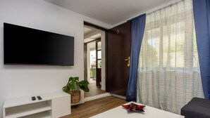 Ett modernt vardagsrum med en platt-TV, ett vitt soffbord, en mörk soffa och ett stort fönster med blå gardiner.