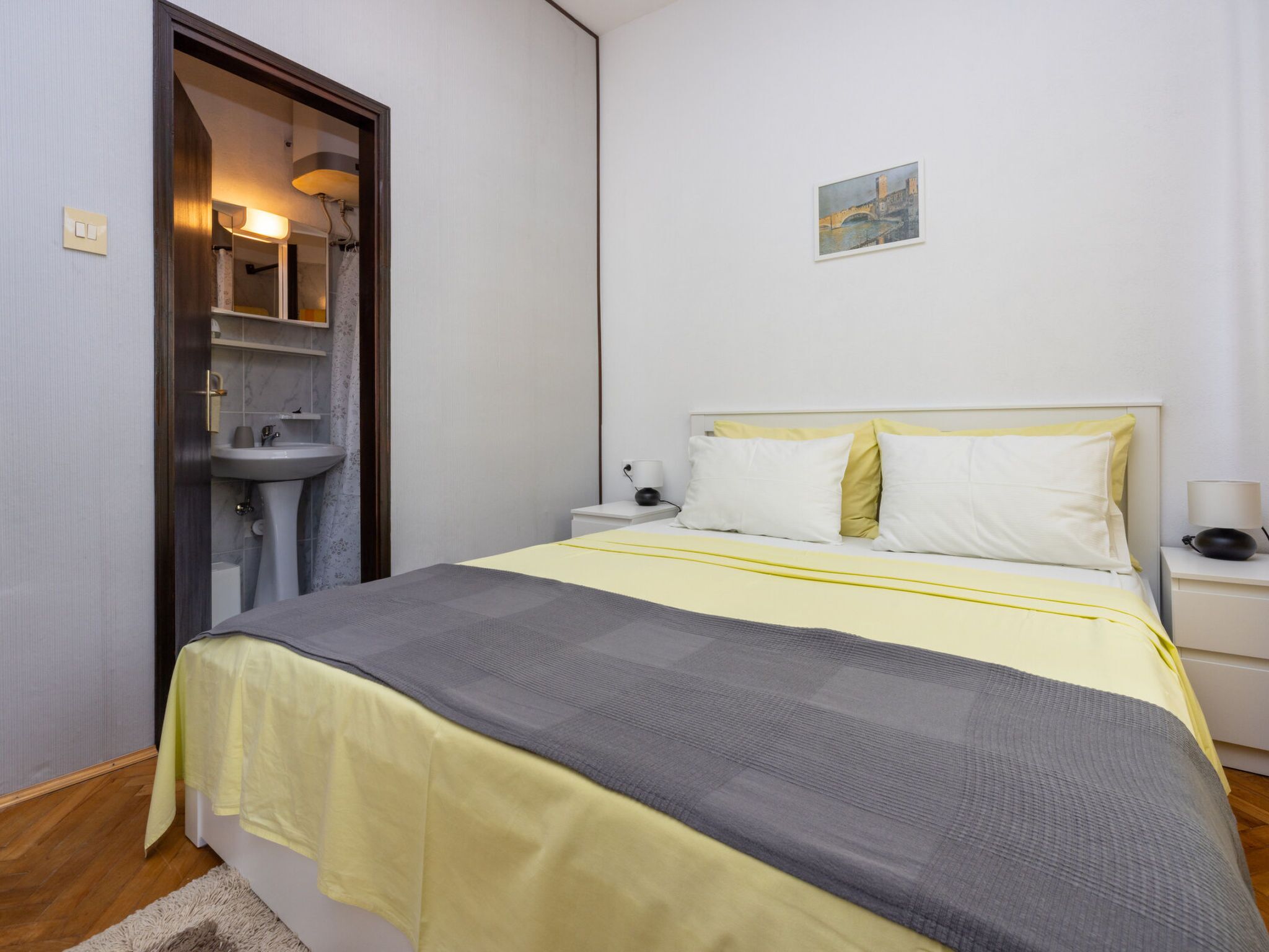 Une chambre d’hôtel avec un lit, des tables de chevet, une petite table, une lampe fixée au mur, un tableau et une salle de bain visible à travers une porte ouverte.