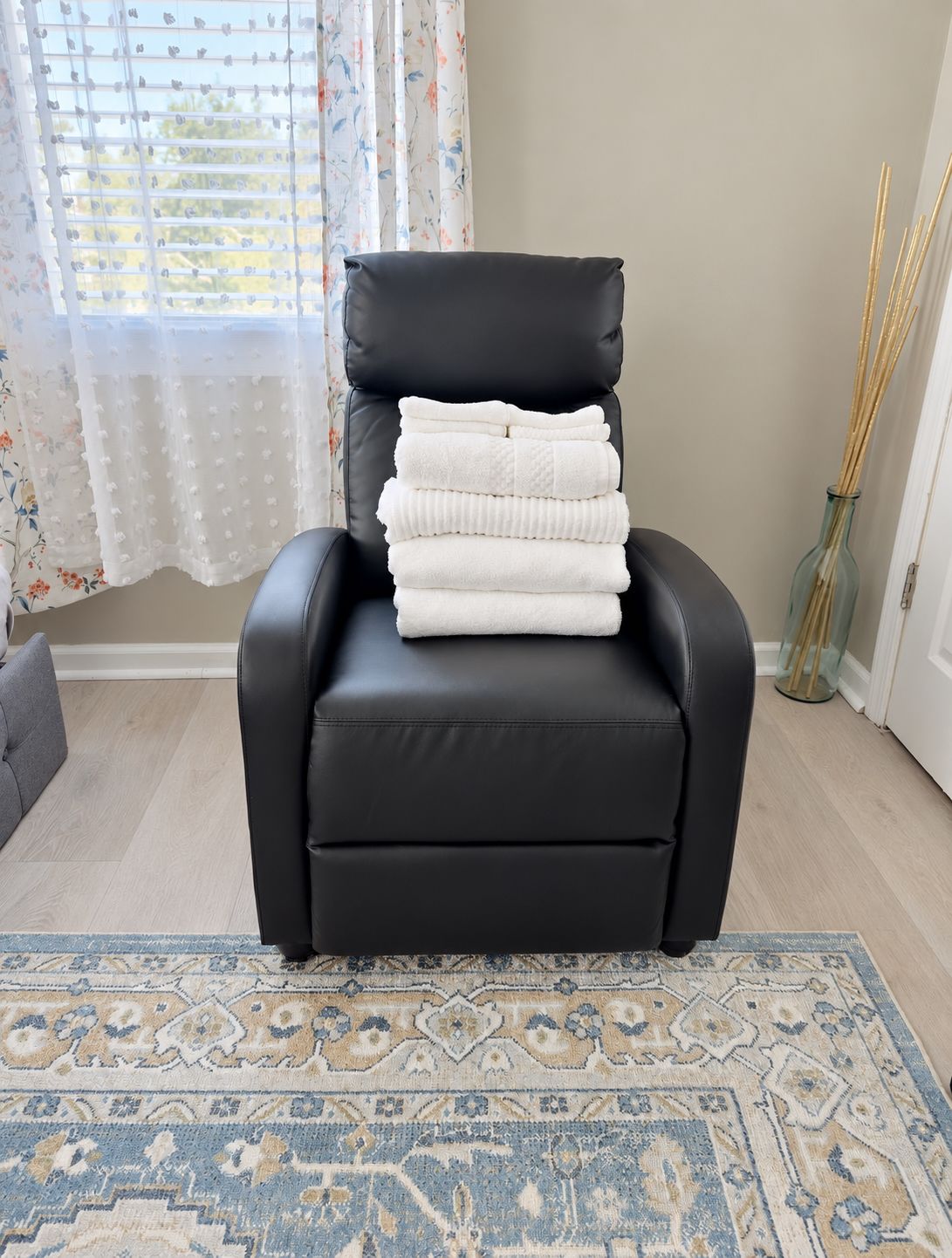 Un fauteuil en cuir noir, avec une pile de serviettes blanches à côté, un tapis à motifs sur le sol et une fenêtre ornée de rideaux à fleurs.