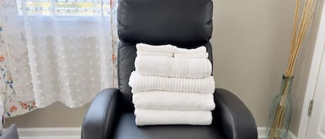 Un fauteuil en cuir noir, avec une pile de serviettes blanches à côté, un tapis à motifs sur le sol et une fenêtre ornée de rideaux à fleurs.