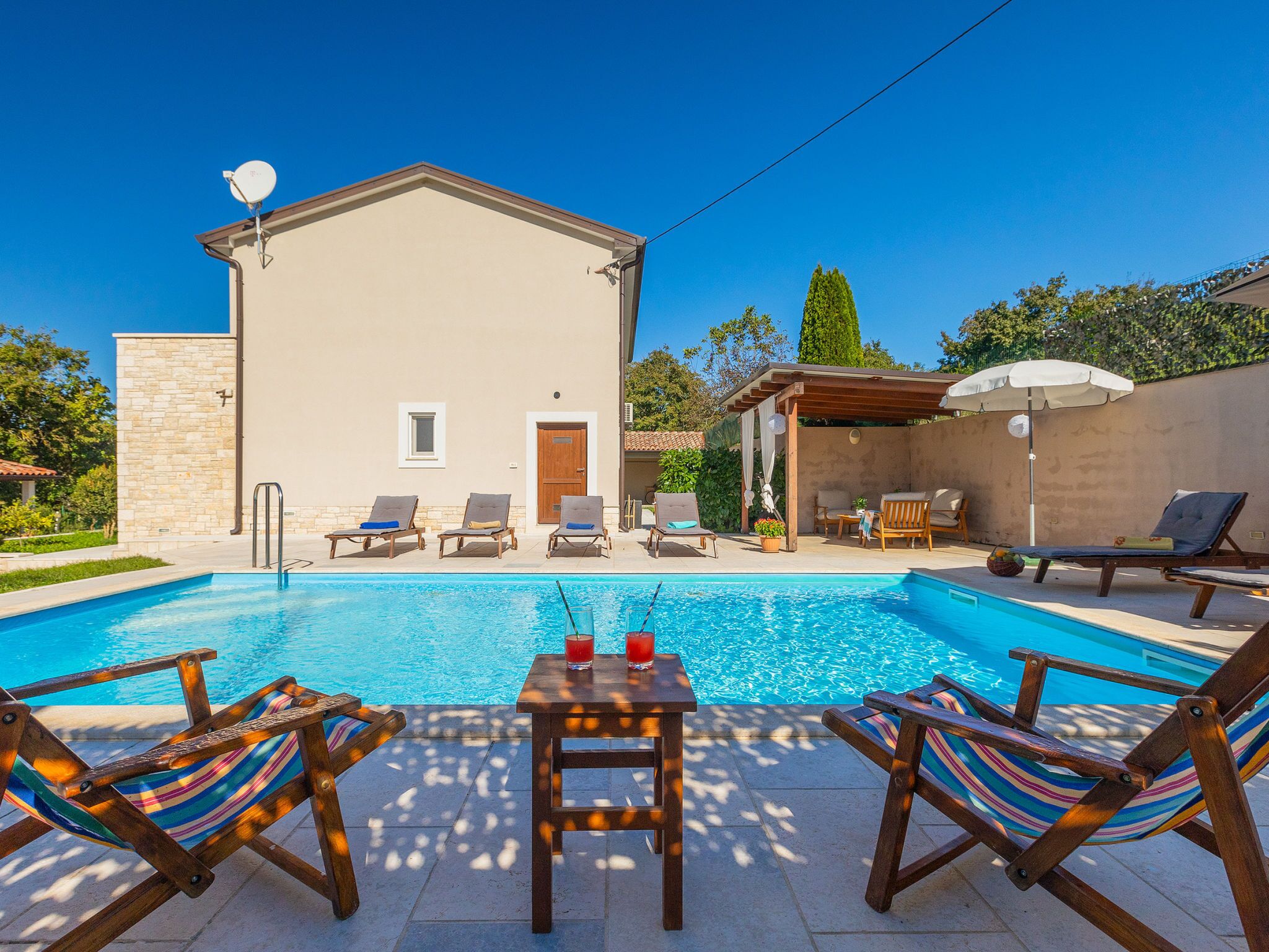 Une piscine avec des chaises longues et une table, un bâtiment avec une porte et une fenêtre, et une pergola avec un parasol.