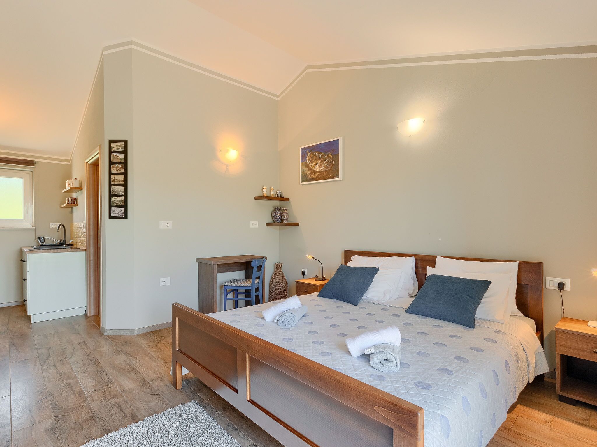 A bedroom with a wooden bed, a bedside table, a nightstand, a small desk with a chair, a window, and a wall-mounted shelf with decorative items.