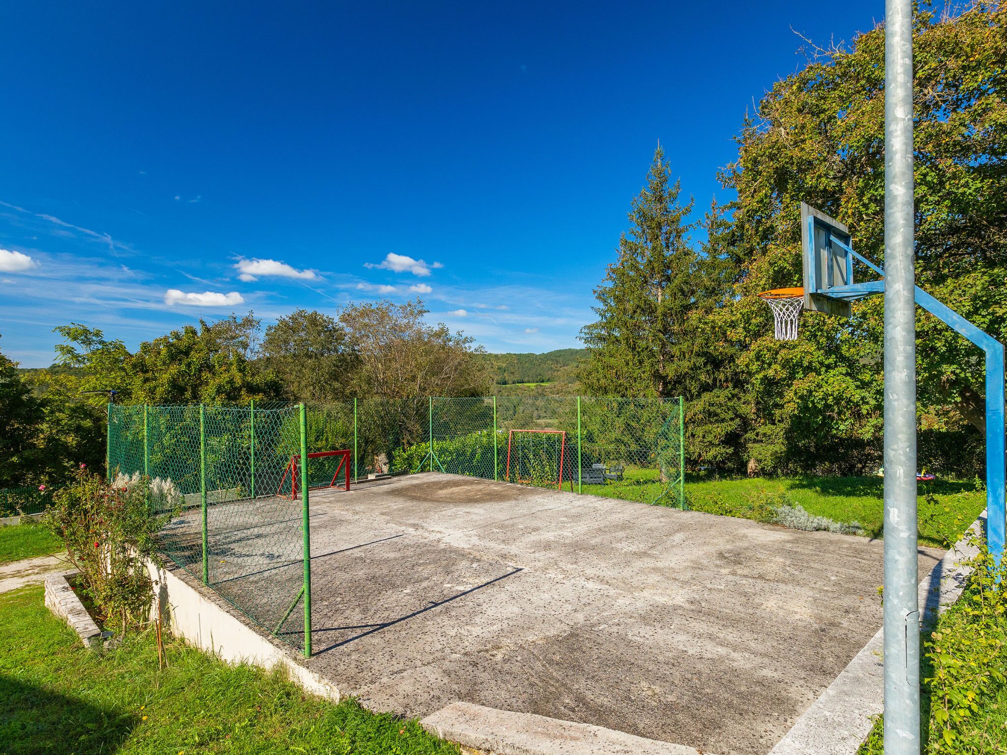 An outdoor basketball court with a chain-link fence, a basketball hoop, and trees in the background.