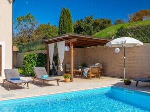 Una piscina con sillas de descanso, una pérgola con mesa y sillas, y vistas a árboles y colinas.