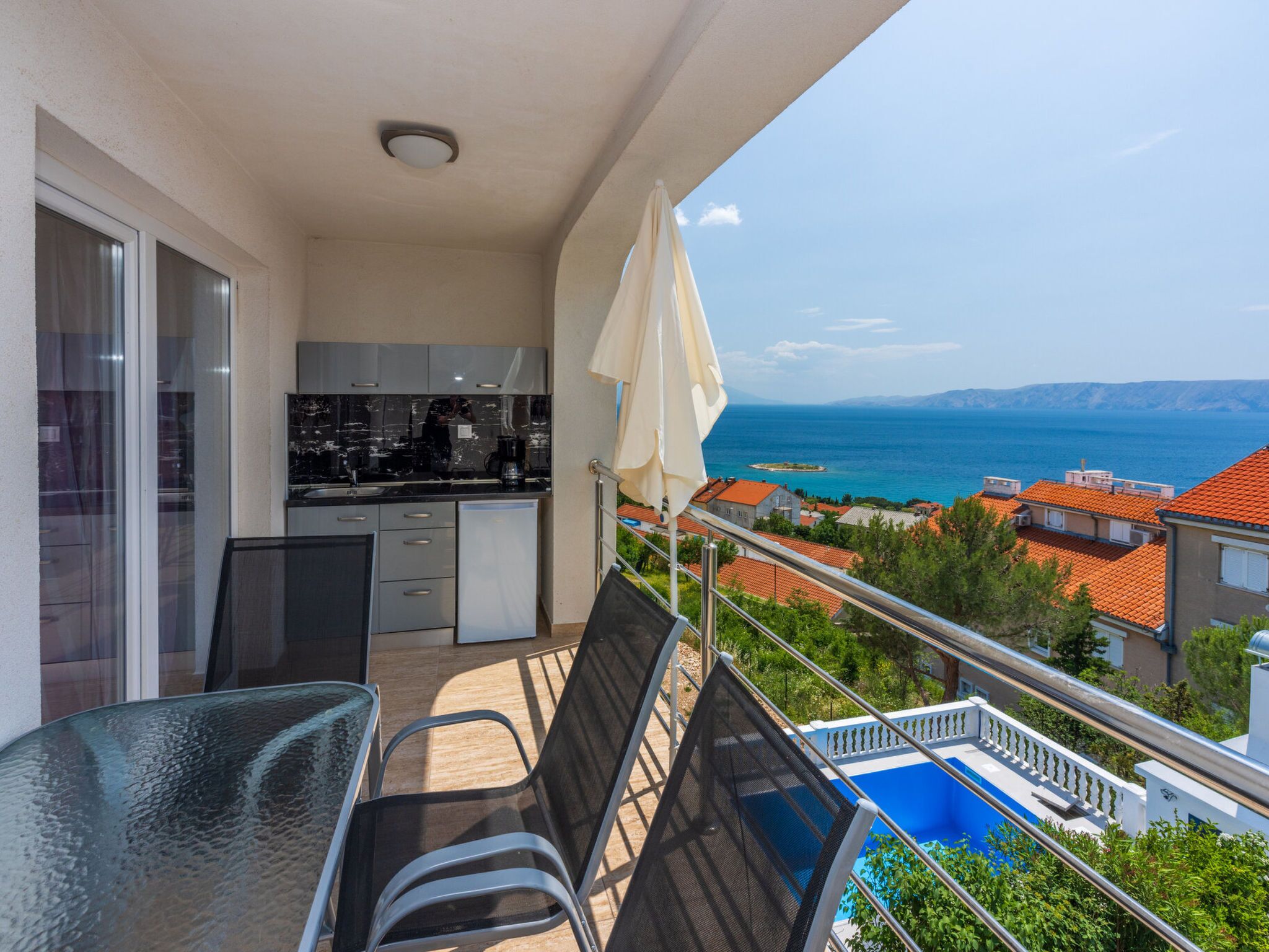 A balcony with a view of the sea, a dining area with chairs, a kitchen, and a pool.