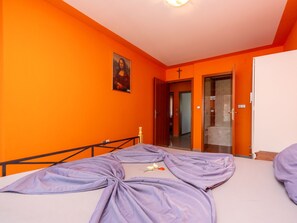 Una habitación con paredes anaranjadas, una cama con colcha morada, un cuadro de una mujer y una puerta que da a un baño.