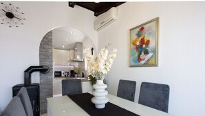 Un comedor con una mesa blanca, un mantel individual negro y un jarrón blanco con flores. Hay sillas tapizadas en gris y un armario negro con un tirador. Una unidad de aire acondicionado está montada en la pared, y un cuadro colorido cuelga sobre la mesa.