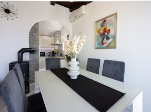 Ein Essbereich mit einem weißen Tisch, einer schwarzen Tischmatte und einer weißen Vase mit Blumen. Graue Sitzmöbel und ein schwarzes Sideboard mit Griff sind ebenfalls vorhanden. Eine Klimaanlage ist an der Wand montiert, und über dem Tisch hängt ein farbenfrohes Gemälde.