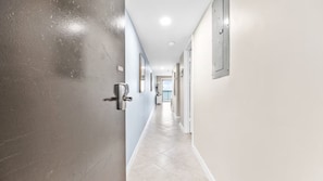 Un couloir avec une porte sur la gauche, un interrupteur sur le mur et un luminaire au plafond.