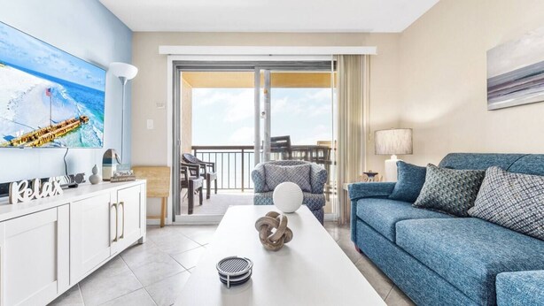 Un salón moderno con un sofá, una mesa de centro y un televisor. Hay un balcón con vistas al océano.