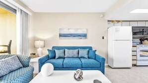 Una sala de estar moderna con un sofá azul, una mesa de centro blanca y una cocina con un refrigerador y estufa blancos.