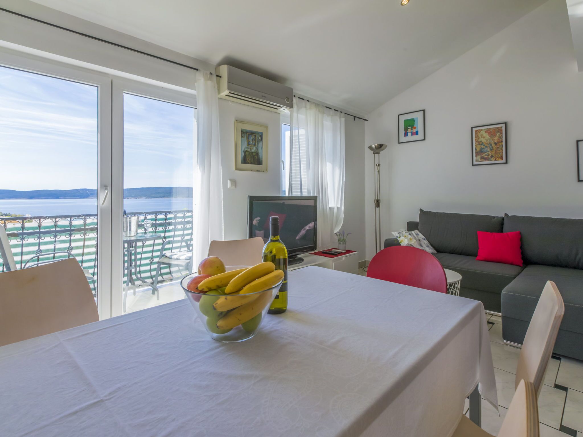 Un salon moderne avec un balcon, une table à manger ornée de fruits, un canapé et des œuvres d’art accrochées aux murs.
