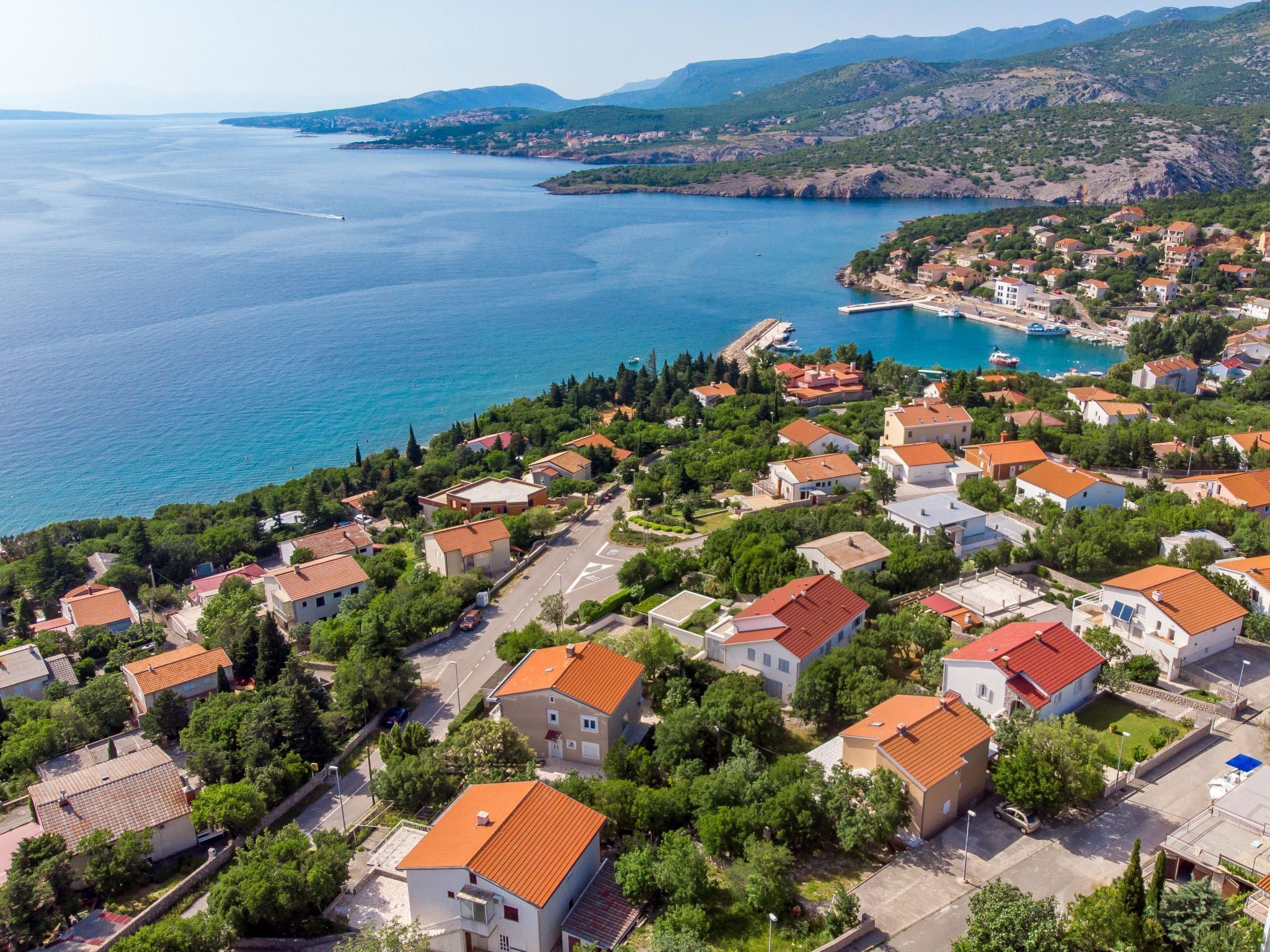 A coastal residential area with houses featuring red-tiled roofs, surrounded by greenery and a view of the sea.