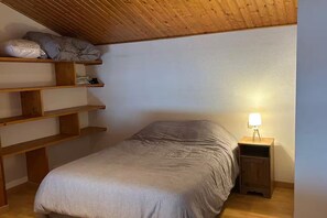 A bedroom with a bed, wooden shelves, a nightstand with a lamp, and a sloped ceiling. - Apartment, breathtaking sea view (bed linen and towels included) (Saint-Gilles-Croix-de-Vie)