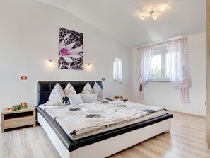 Ein Schlafzimmer mit einem Bett, einem Nachttisch, einer Wandleuchte, einem Fenster mit Vorhängen und einem an der Wand befestigten Spiegel.