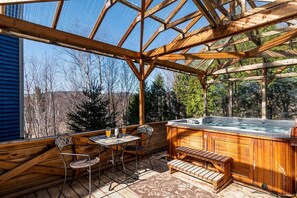 Eine Holzterrasse mit Whirlpool, Tisch und Stühlen sowie Blick auf Bäume und Hügel.