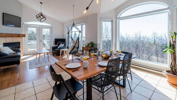 A modern dining area with a wooden table, black chairs, and a view of trees through large windows. - Bleuzen • Vue splendide • 2 chambres • Foyer • Balcon (Petite-Rivière-Saint-François)