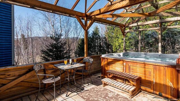 A wooden deck with a hot tub, a table with chairs, and a view of trees and hills. - Bleuzen Charlevoix • 3 chambres • 2 salle-de-bain • SPA privé • Foyer • Terrasse (Petite-Rivière-Saint-François)