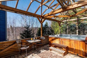 A wooden deck with a hot tub, a table with chairs, and a view of trees and hills. - Bleuzen Charlevoix • 3 chambres • 2 salle-de-bain • SPA privé • Foyer • Terrasse (Petite-Rivière-Saint-François)