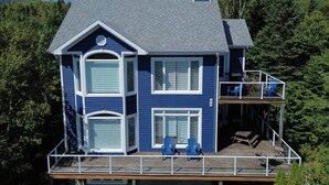 A two-story house with a balcony, large windows, and a deck with outdoor furniture. - Bleuzen Charlevoix • 3 chambres • 2 salle-de-bain • SPA privé • Foyer • Terrasse (Petite-Rivière-Saint-François)