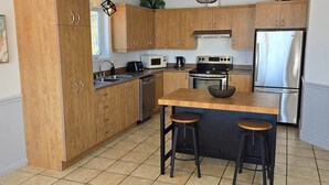 A modern kitchen with wooden cabinets, stainless steel appliances, and a central island with stools. - Bleuzen Charlevoix • 3 chambres • 2 salle-de-bain • SPA privé • Foyer • Terrasse (Petite-Rivière-Saint-François)