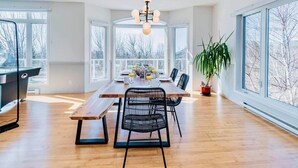 A spacious dining area with a wooden table, black chairs, and a large window offering a view of trees. - Bleuzen Charlevoix • 3 chambres • 2 salle-de-bain • SPA privé • Foyer • Terrasse (Petite-Rivière-Saint-François)