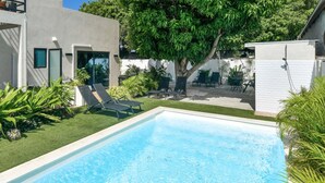A rectangular swimming pool with clear blue water, surrounded by a well-maintained lawn and several lounge chairs. - Mango Gardens - Tropical Living Made Simple (Oranjestad)