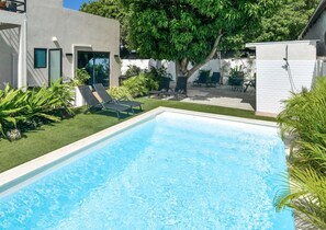 A rectangular swimming pool with clear blue water, surrounded by a well-maintained lawn and several lounge chairs. - Mango Gardens - Tropical Living Made Simple (Oranjestad)