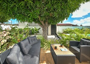 An outdoor patio with wicker furniture, a wooden table, and a large tree providing shade. - Mango Gardens - Tropical Living Made Simple (Oranjestad)