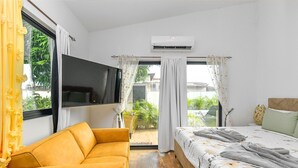 A bedroom with a sofa, a bed with a floral duvet, an air conditioning unit, and a TV mounted on the wall. - Mango Gardens - Tropical Living Made Simple (Oranjestad)