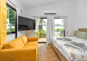 A bedroom with a sofa, a bed with a floral duvet, an air conditioning unit, and a TV mounted on the wall. - Mango Gardens - Tropical Living Made Simple (Oranjestad)