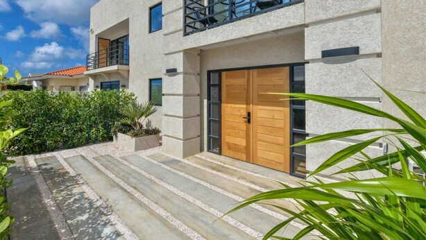 A modern two-story building with a balcony, a wooden door, and a gravel pathway leading to the entrance. - Mango Gardens - Tropical Living Made Simple (Oranjestad)
