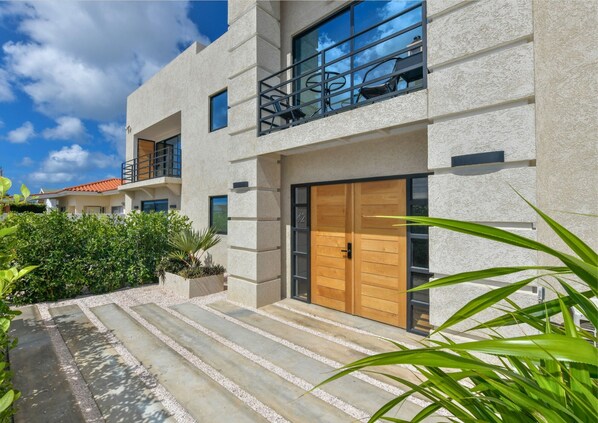 A modern two-story building with a balcony, a wooden door, and a gravel pathway leading to the entrance. - Mango Gardens - Tropical Living Made Simple (Oranjestad)