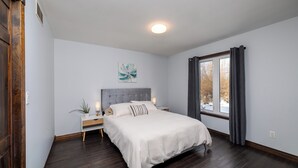A bedroom with a large bed, a nightstand, a wall-mounted light, a window with curtains, and a ceiling light. - The Bognor House (Meaford)
