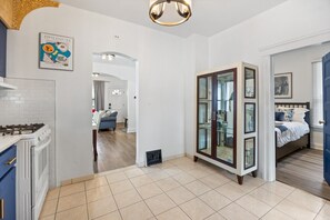 A modern kitchen with a gas stove, a dining area with a blue sofa, and a bedroom with a bed and window blinds. - Little Italy 4BR Home | Garage, Yard & Parking (Windsor)