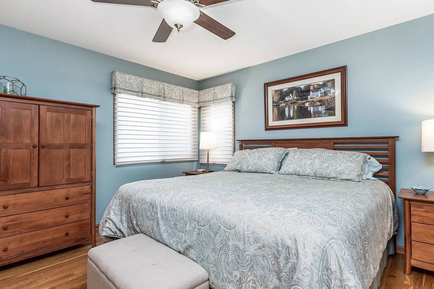 A bedroom with a bed, a nightstand, a wooden wardrobe, a window with blinds, and a ceiling fan.