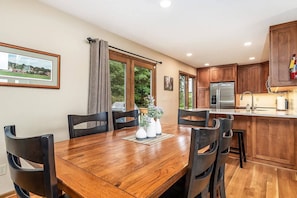 A dining area with a wooden table and chairs, a kitchen with wooden cabinets, and a large window with curtains. - Beautiful Peaceful Galena Home. Hot Tub, Sunroom, Pool Table, Arcade  4BR, 3.5BA (Galena)
