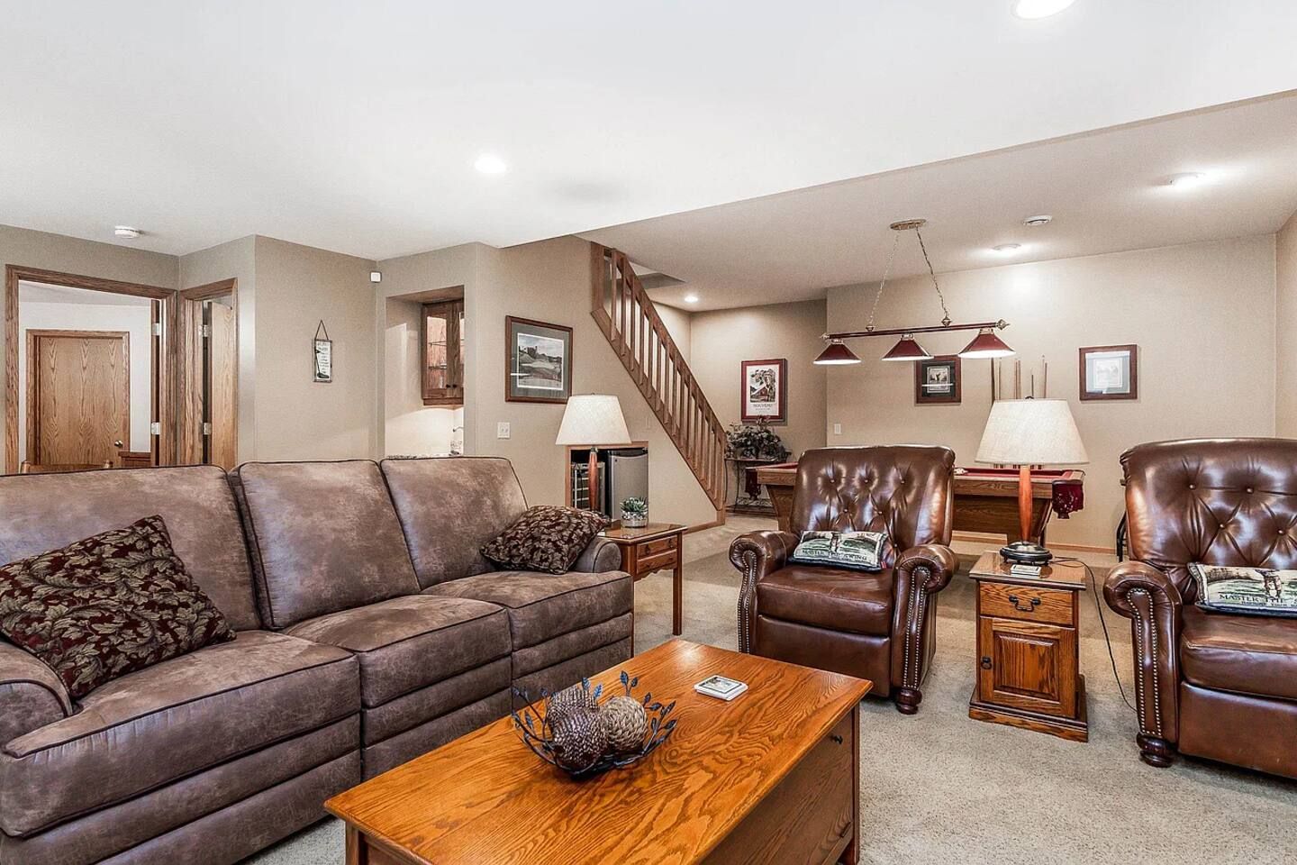 A living room with a brown leather sofa, a wooden coffee table, and two leather armchairs. A staircase is visible in the background.