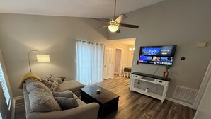 Uma sala de estar com sofá, mesa de centro, TV e ventilador de teto.